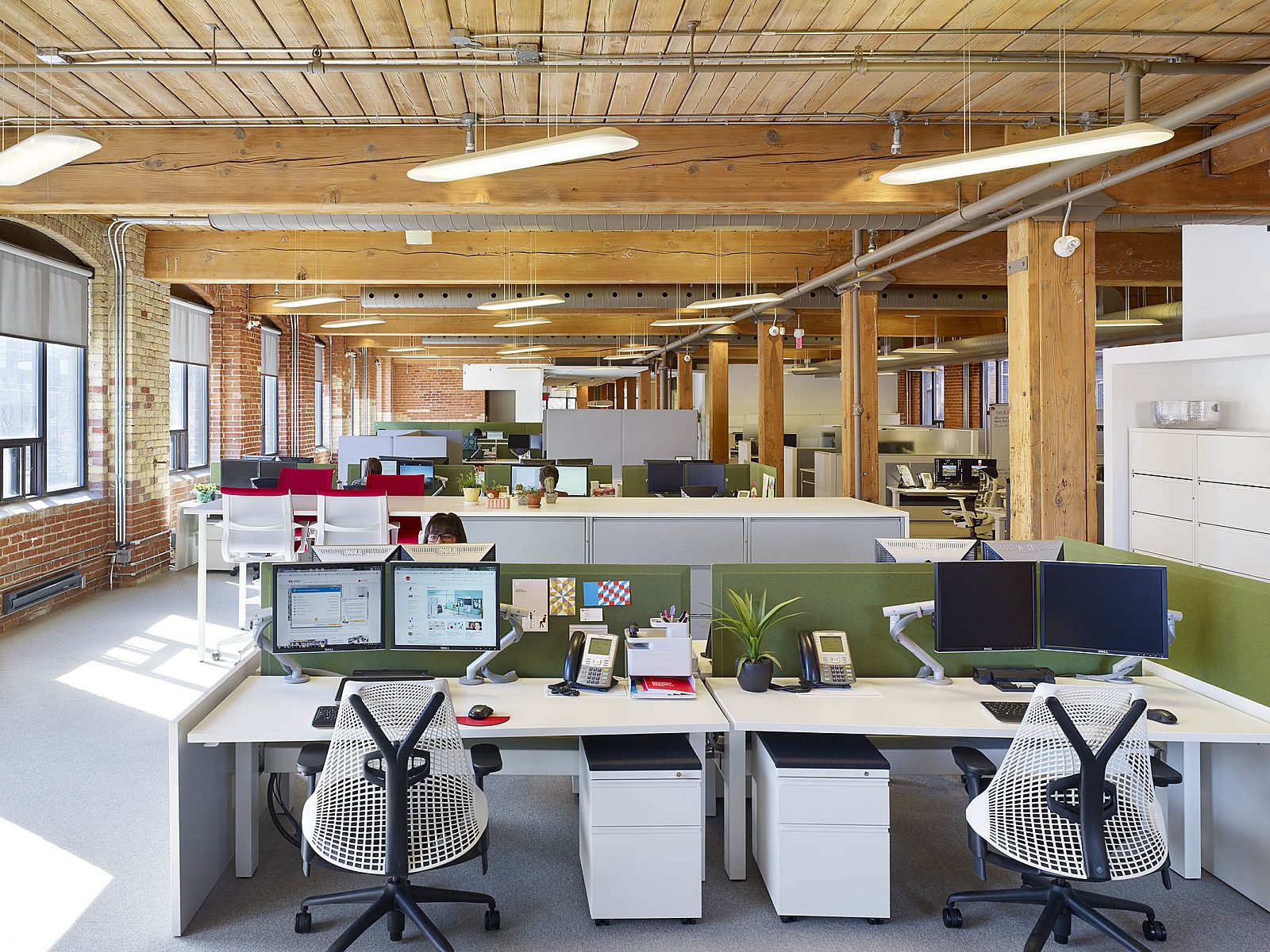 Modern open-plan office with exposed brick walls large windows. Bright desks feature computers, phones, and plants, creating a lively work atmosphere.